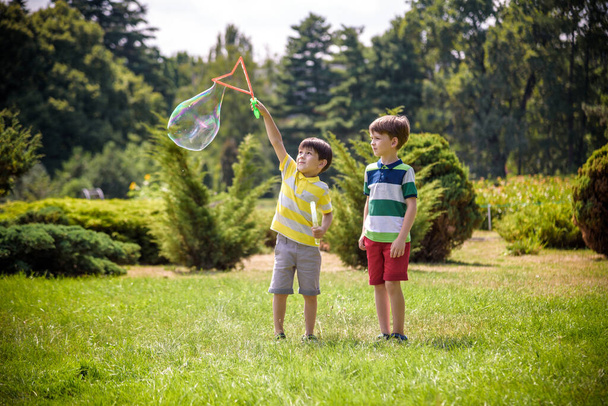Мальчик надувает мыльные пузыри, в то время как возбужденный ребенок наслаждается пузырьками. Счастливый подросток и его брат в парке наслаждаются созданием мыльных пузырей. Концепция счастливого детства. - Фото, изображение