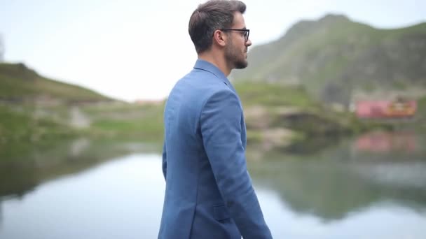 δροσερό έξυπνο casual επιχειρηματίας περπάτημα κοντά σε μια λίμνη στα βουνά, βάζοντας τα χέρια του στην τσέπη, θαυμάζοντας τη θέα, πηγαίνει προς τα κάτω και τον καθορισμό μανίκι του, στη συνέχεια, ανεβαίνει - Πλάνα, βίντεο