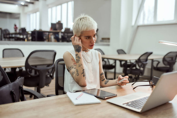 Appel vidéo. Jeune femme d'affaires tatouée blonde élégante portant des écouteurs sans fil tout en étant assise à sa place de travail dans le bureau moderne - Photo, image