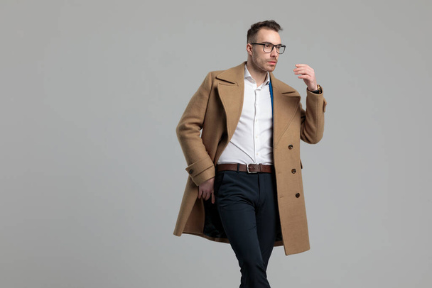 confident young fashion model holding hand in fashion pose, wearing glasses and long coat, looking to side and standing on grey background - Photo, Image