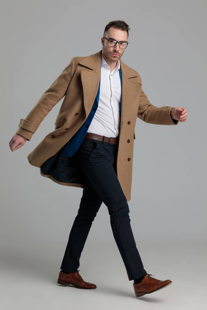 ελκυστική smartcasual τύπος φορώντας μακρύ παλτό με τα πόδια σε γκρι φόντο - Φωτογραφία, εικόνα