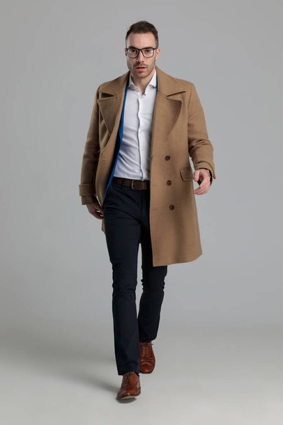 sexy smartcasual guy wearing long coat walking on grey background - Fotó, kép