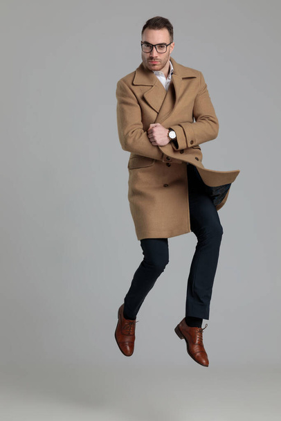 ελκυστικός νεαρός με μακρύ παλτό και άλμα σε γκρι φόντο - Φωτογραφία, εικόνα