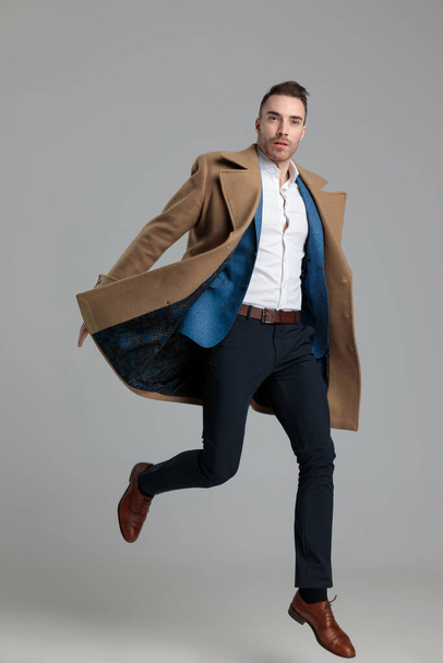 attractive young businessman in longcoat jumping on grey background - Foto, Imagem