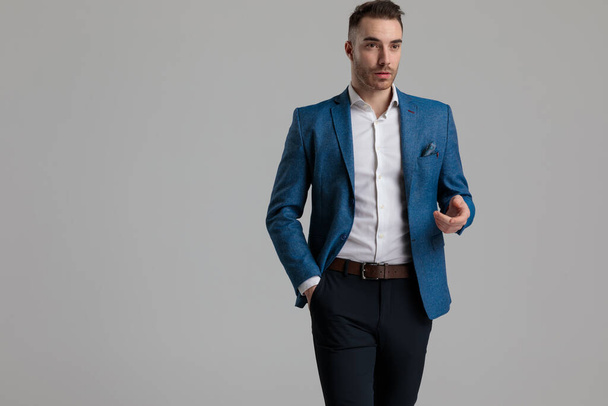 sexy smarter Mann hält Hand in Mode-Pose, grüßt und geht auf grauem Hintergrund - Foto, Bild