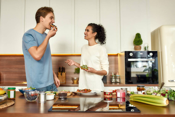 We are what we eat. Happy couple, vegetarians cooking breakfast in the kitchen. Young man and woman tasting sandwiches that they made together. Vegetarianism, healthy food, diet, stay home concept - Photo, image