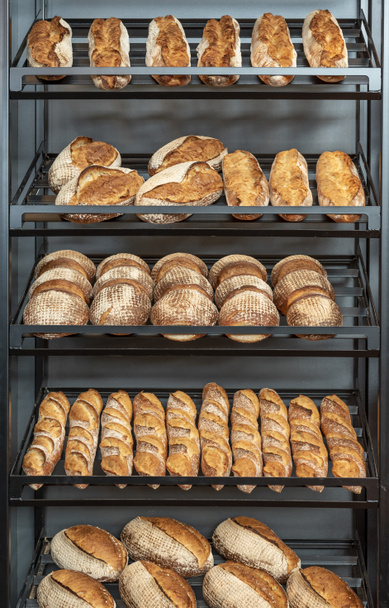 Pan recién horneado en un estante de metal dentro de una panadería. Varios tipos y tamaños de pan blanco
. - Foto, Imagen