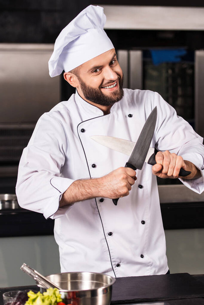 Profesjonalny kucharz pozujący w kuchni restauracji. Uśmiechnięty kucharz ze skrzyżowanymi nożami. - Zdjęcie, obraz