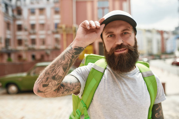 So fast, So easy. Brutal bearded delivery man in cap looking at camera, standing with backpack outdoors, ready to work. Courier, delivery service concept - Фото, зображення