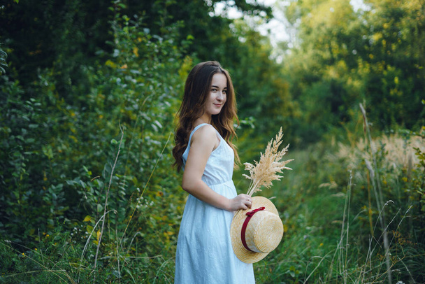 Mooi meisje in een witte jurk met een boeket gedroogde bloemen. Jonge vrouw in een groen park. Meisje in een jurk en een zomerse zonnige dag - Foto, afbeelding