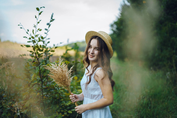 Smiling girl in a straw hat collects flowers. Girl in a summer dress in the tall grass. Woman in a green field with a bouquet of dried flowers - Foto, afbeelding