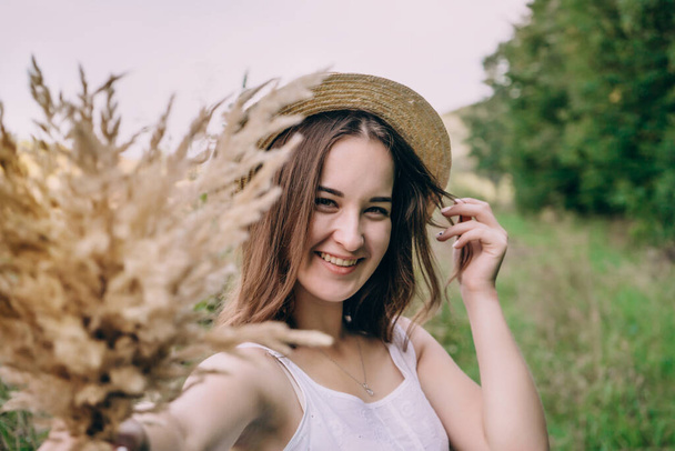Happy young woman in a white dress in a straw hat. Smiling girl with a bouquet of dried flowers. Girl walks in a green park on a spring day - Фото, зображення