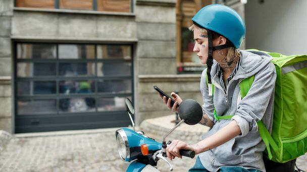 Mujer joven en casco con mochila térmica usando teléfono inteligente mientras entrega comida en un scooter
 - Foto, Imagen
