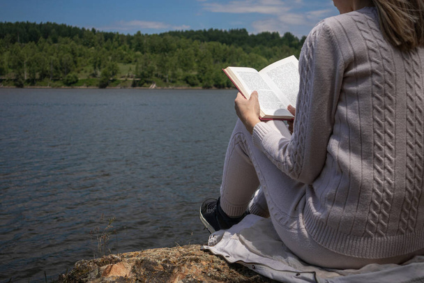 自然界の湖の近くに座って読んで足の本を持つ少女。セーターのブロンドの女性は、湖の海岸に大きな岩の上に座っている。リラクゼーションの概念。閉じる、選択的フォーカス - 写真・画像