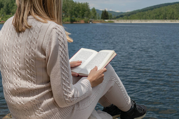 晴れた日にはセーターを着て湖の水に近い本を読んでいる少女。ブロンドの女の子の足に本を湖の岸に大きな岩の上に座っている。リラクゼーションの概念。閉じる、選択的フォーカス - 写真・画像