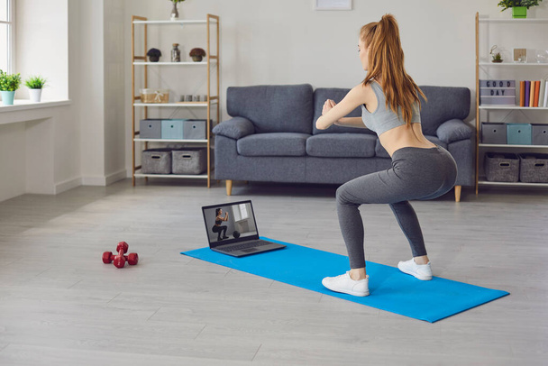 Online sports. Red haired girl watching video training on laptop computer and doing squats in living room - Fotó, kép