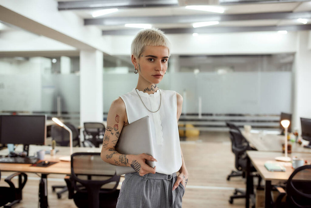 I am ready to work. Young, stylish and attractive tattooed business woman holding her laptop and looking at camera while standing in the modern coworking space - Фото, зображення