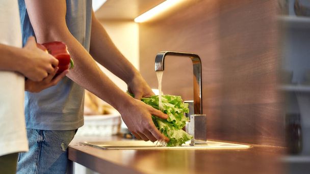 Man s hands washing lettuce in modern kitchen sink before cooking, preparing a meal. Woman helping him, holding other vegetables. Vegetarianism, healthy food, hygiene concept - Foto, afbeelding