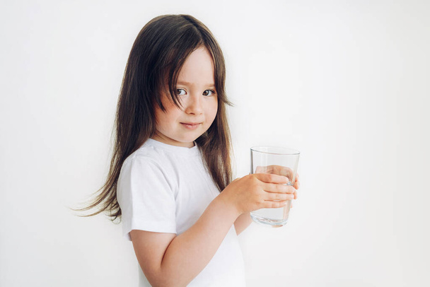 Ένα παιδί με λευκό μπλουζάκι πίνει νερό. Το κοριτσάκι στέκεται με ένα ποτήρι νερό. - Φωτογραφία, εικόνα