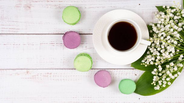 Kleurrijke macarons, een kop koffie en lelies van de vallei op houten achtergrond, close-up plat gelegd. Concept van ontbijt en lente tijd. Kopieerruimte. - Foto, afbeelding