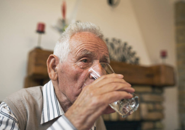 老人は家のテーブルでガラスから水を飲む。高齢者における水分補給の概念 - 写真・画像