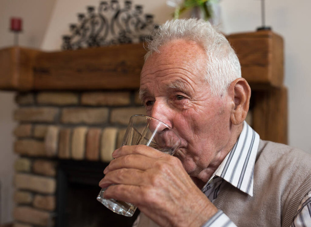 老人は家のテーブルでガラスから水を飲む。高齢者における水分補給の概念 - 写真・画像