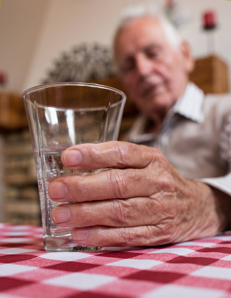 Close up of old man's hand holding glass of water at table - Foto, afbeelding