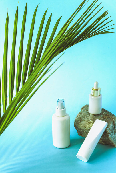 Kosmetiktonikum, Cremetubus und Serum in leeren Packungen auf blauem Hintergrund mit Palmblättern. Konzept natürlicher Hautpflegeprodukte. - Foto, Bild