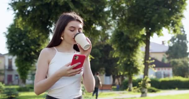 Girl Wearing Walking on the Street scrolls and reads amusing article on her Modern Smartphone. Holding Cup in her left hand. Beautiful lady is busy on her phone. - Filmagem, Vídeo