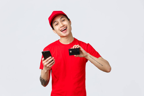 Freundlicher Zusteller in rotem einheitlichen T-Shirt und Mütze, Smartphone-Bildschirm und Kreditkarte vorzeigend, empfiehlt Anwendung zur Verfolgung von Online-Shopping-Bestellungen. Kurier wirbt für Carrier-Service - Foto, Bild