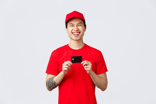 Vrolijke glimlachende Aziatische bezorger in rode pet en t-shirt uniform, lachen als het bevorderen van gemakkelijke betaling voor online bestellingen terwijl op coronavirus quarantaine thuis blijven, koerier houden credit card - Foto, afbeelding