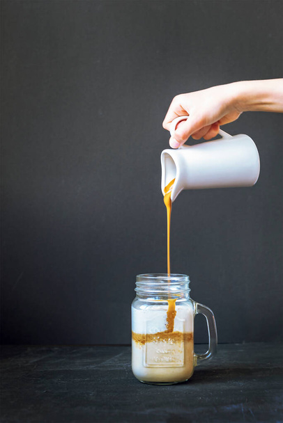 Kaffeeschaum wird in einen Becher mit Milch gegossen. Dalgona Kaffee kochen. Schwarzer Hintergrund, Nahaufnahme - Foto, Bild