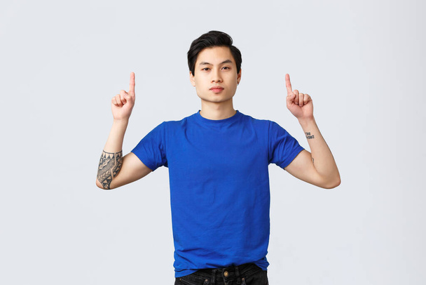 Unterschiedliche Emotionen, Menschen Lebensstil und Werbekonzept. Selbstbewusster, seriöser asiatischer Mann in blauem T-Shirt mit Tätowierungen, Zeigefinger nach oben, informiert Kunden, zeigt Weg zum Banner, grauer Hintergrund - Foto, Bild