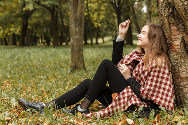 Beautiful romantic girl in a park autumn scenery looking at the camera, sitting down next to a tree, enjoying the perfect weather in a sunny day. Gorgeous young woman outdoors. Full length body shot - Photo, image