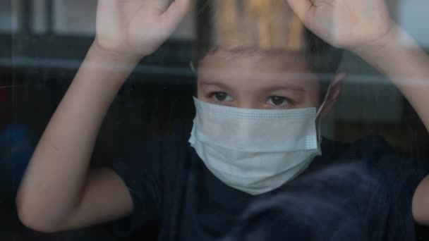 Young boy in a medical mask looks out the window. Self-isolation in quarantine, coronavirus, covid 19. - Кадри, відео
