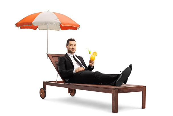 Man in a black suit lying on a sunbed with umbrella and drinking a cocktail isolated on white background - Fotografie, Obrázek