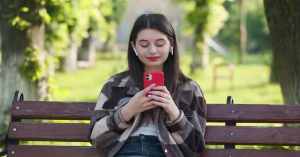 Attractive woman browsing on mobile phone in public park. City, urban background. Girl listening to music in the park using mobile phone and wearing headphones. - Felvétel, videó