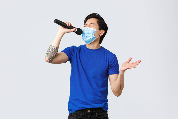 Covid-19 lifestyle, people emotions and leisure on quarantine concept. Handsome funny young male student staying home during coronavirus pandemic, wear mask and singing karaoke in microphone - Photo, Image