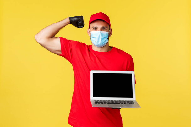 covid-19, safe shopping, express delivery and order tracking concept. Strong handsome courier in red uniform and medical mask, flex biceps and showing laptop screen, smiling - Photo, Image