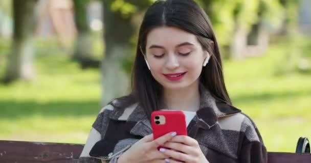 Woman enjoying music and Typing Messages in the park. Joyful young woman listening to music wear earphone looking at smartphone smile at caucasian urban background - Filmagem, Vídeo