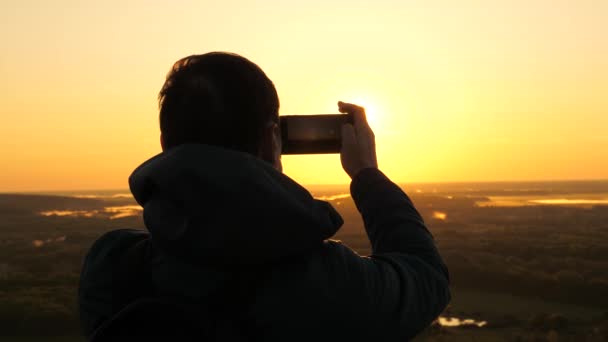 the traveler takes photos and videos on his smartphone at dawn, from the mountain in rays of the beautiful sun. free peasant tourist enjoys a beautiful view of nature from a high hill. travel concept - Footage, Video