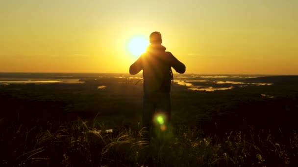 a traveler with a backpack stands on edge of mountain in rays of dawn, raises his hands in air and enjoys the victory, beautiful sun and landscape. free male tourist travels alone in park - Footage, Video