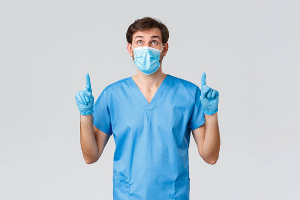 Covid-19, healthcare workers and hospital concept. Doctor reading information on top advertisment banner, wearing scrubs and medical mask, gloves to prevent getting coronavirus, point up - Photo, Image