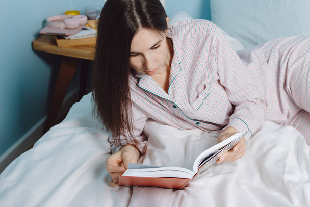 Young calm brunette millennial cheerful mixed race woman student relaxing reading book lying on bed with white sheets - Fotografie, Obrázek