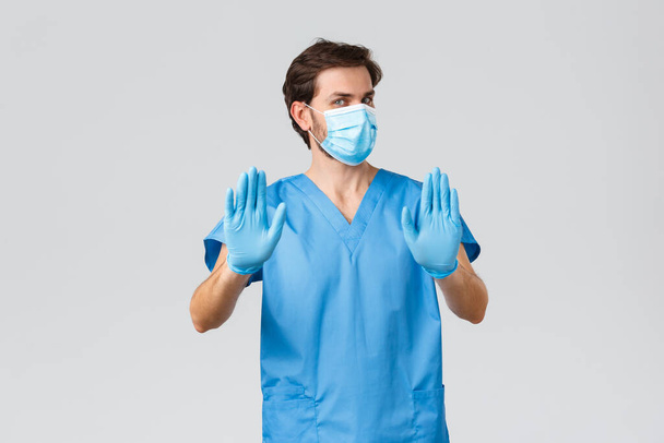 Coronavirus outbreak, healthcare workers fighting disease, hospitals concept. Reluctant and displeased doctor in medical mask, gloves and scrubs, show stop, refuse or reject offer, waving hands - Photo, Image