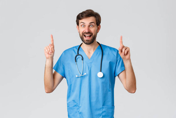 Trabajadores de la salud, pandemia y concepto de brote de coronavirus. Entusiasta médico barbudo sonriente en uniformes azules, estetoscopio, señalando los dedos hacia arriba, mostrar el anuncio superior, se ven sorprendidos y felices
 - Foto, Imagen