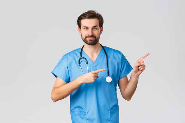 Healthcare workers, pandemic and coronavirus outbreak concept. Professional doctor, physician or nurse in blue scrubs, stethoscope, pointing fingers right and smiling, recommend promo - Photo, Image