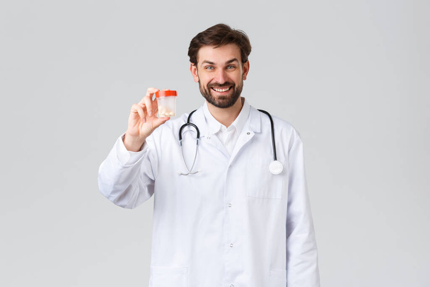 Hospital, healthcare workers, covid-19 treatment concept. Handsome smiling doctor in white scrubs with stethoscope, showing pills, advice patient presctiption, show medication, drugstore promo - Photo, Image