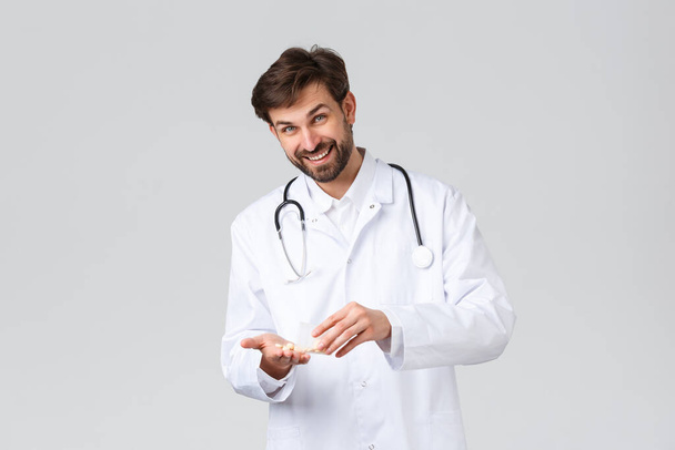 Hospital, healthcare workers, covid-19 treatment concept. Handsome bearded doctor in white scrubs with stethoscope, showing drugs or pills presribed to patient, smile camera, grey background - Fotoğraf, Görsel