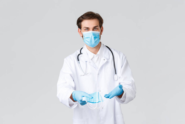 Hospital, healthcare workers, covid-19 treatment concept. Doctor in personal protective equipment and scrubs, holding medical masks, showing patients methods of protecting health during coronavirus - Photo, Image